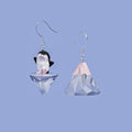 Mismatched Arctic Drift earrings featuring a geometric penguin standing on a floating iceberg and a separate faceted iceberg charm, worn on silver hooks.