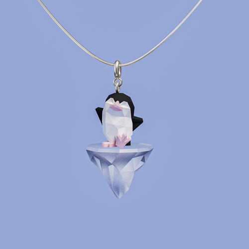 Arctic Drift necklace with a geometric penguin standing on a faceted floating iceberg pendant, hanging from a silver chain against a soft blue background.