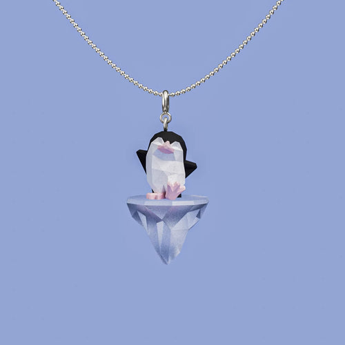 Arctic Drift necklace with a geometric penguin standing on a faceted floating iceberg pendant, hanging from a silver chain against a soft blue background.